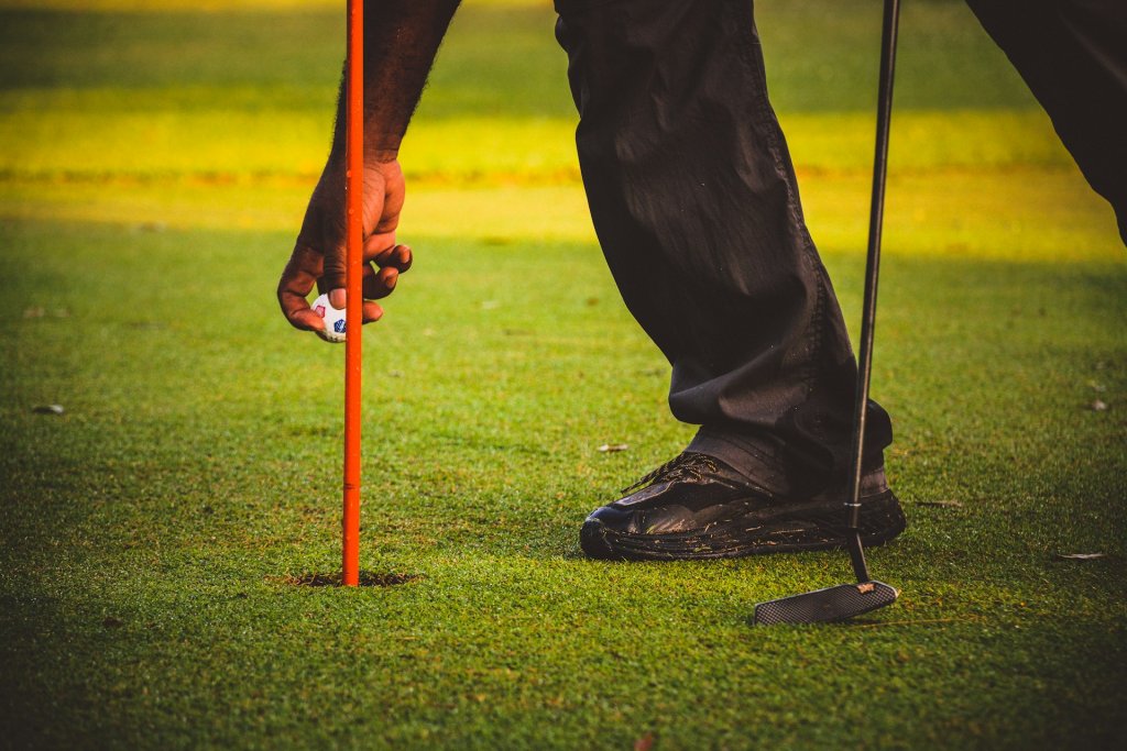 A golfer holding a club in one hand while retrieving his ball from the cup next to the flagstick.