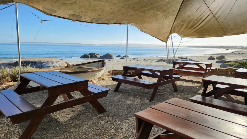 Wooden benches and outdoor tables under a shade canopy overlooking the ocean in Paternoster, South Africa.