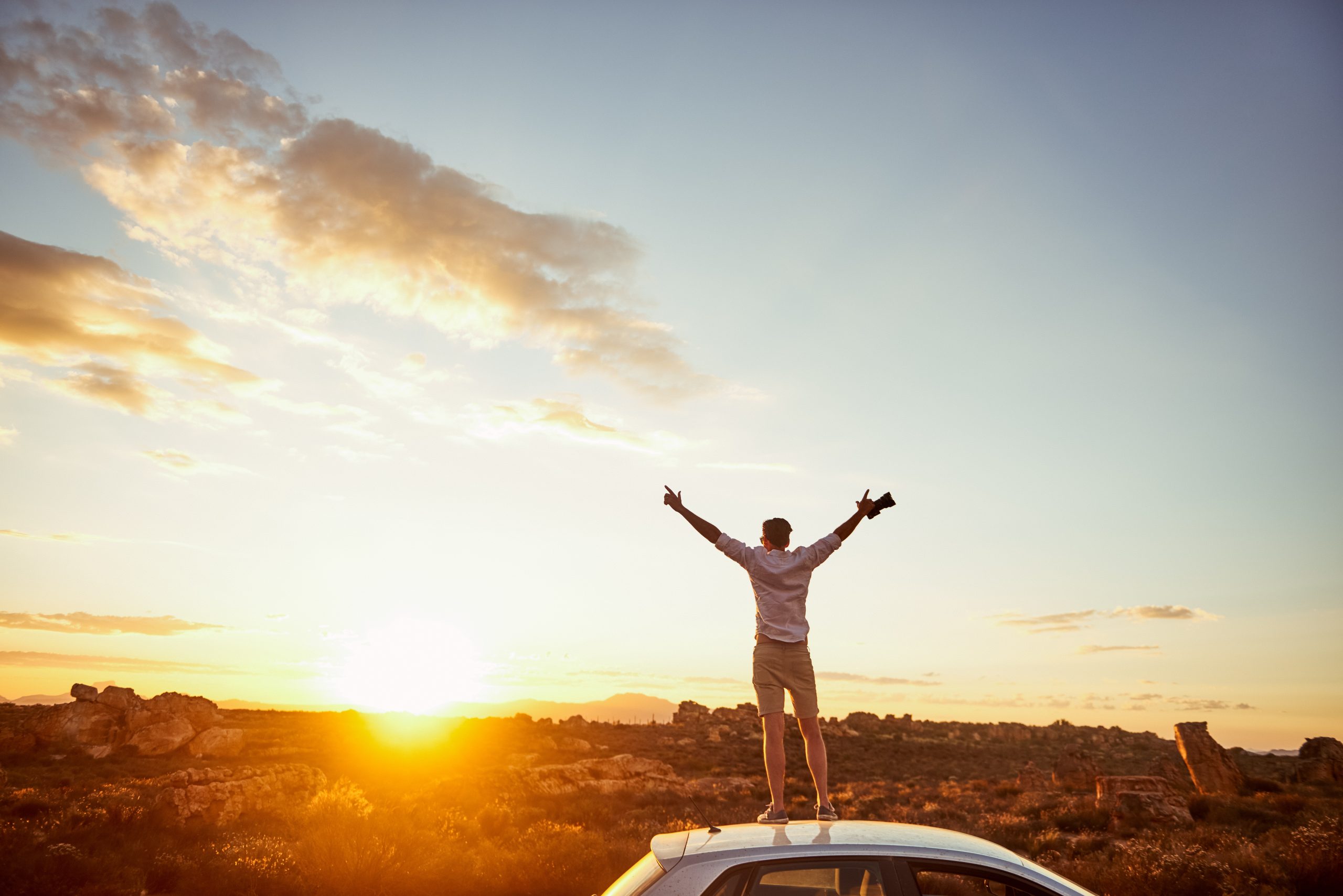 Rearview shot of a young man standing on top of a car with his arms outstretched in a rural landscape