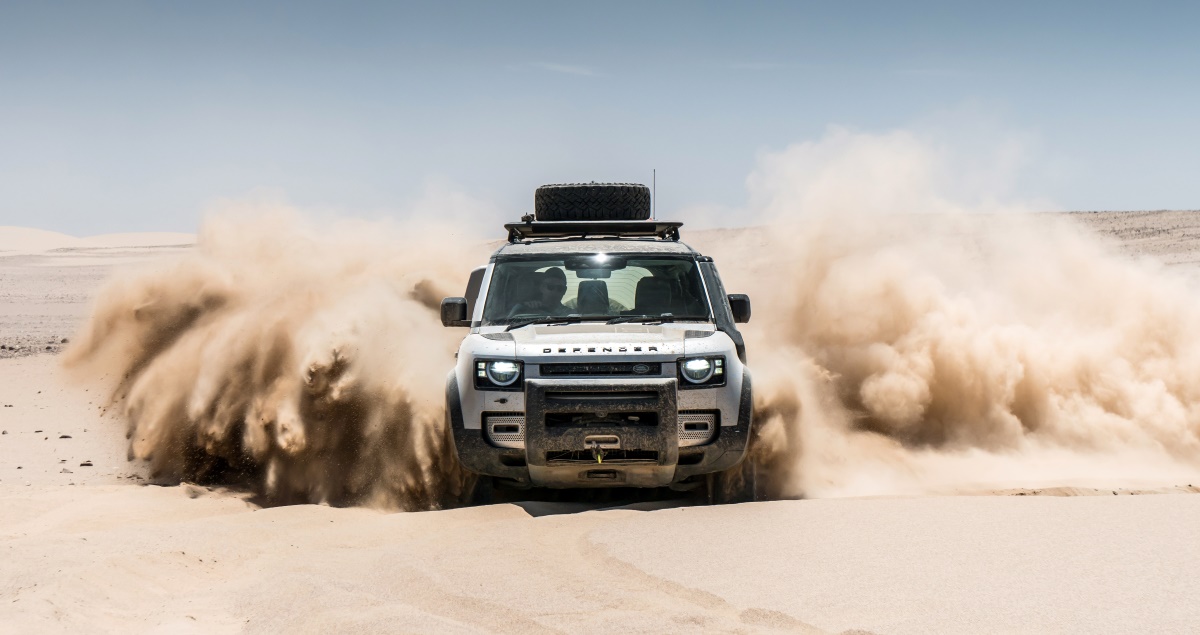 A Land Rover Defender drives through a sand dune.