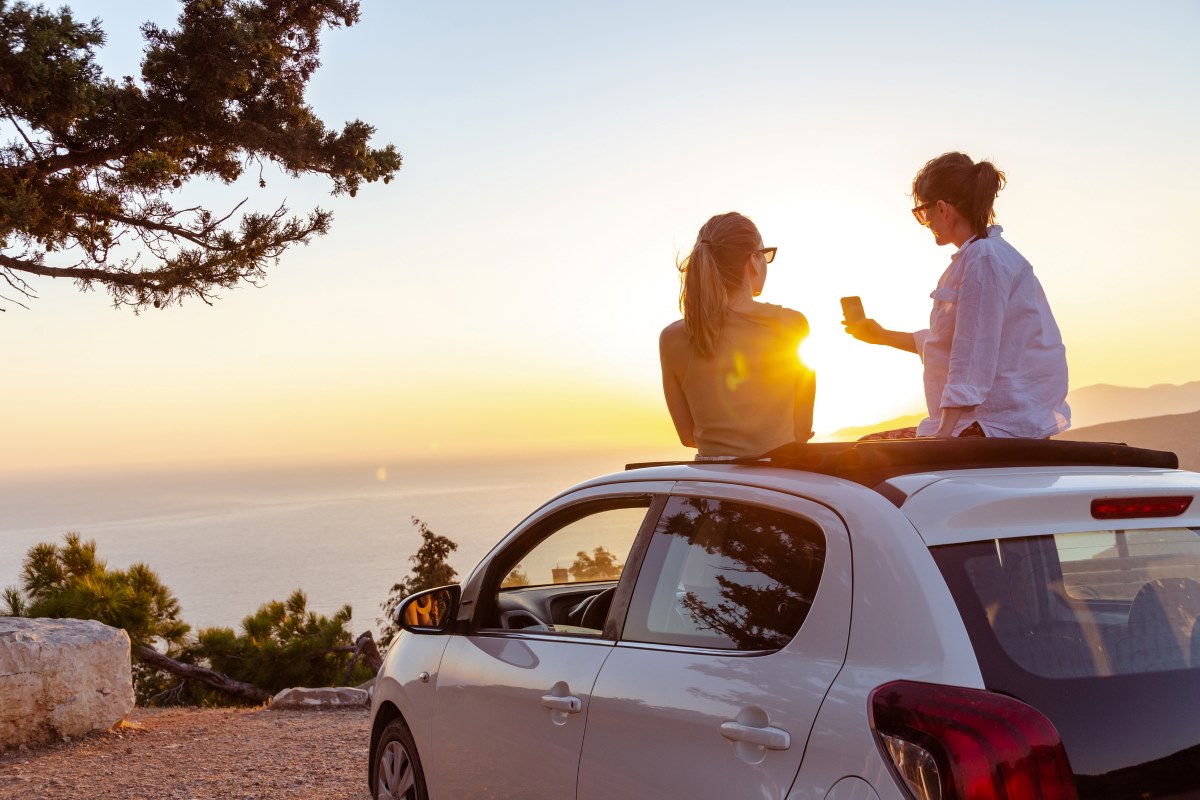 Two women sit on the roof of a car at sunset.