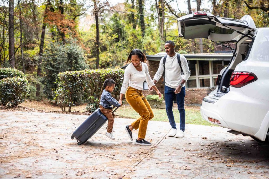 Mother pulling young boy on suitcase and loading car for family vacation.