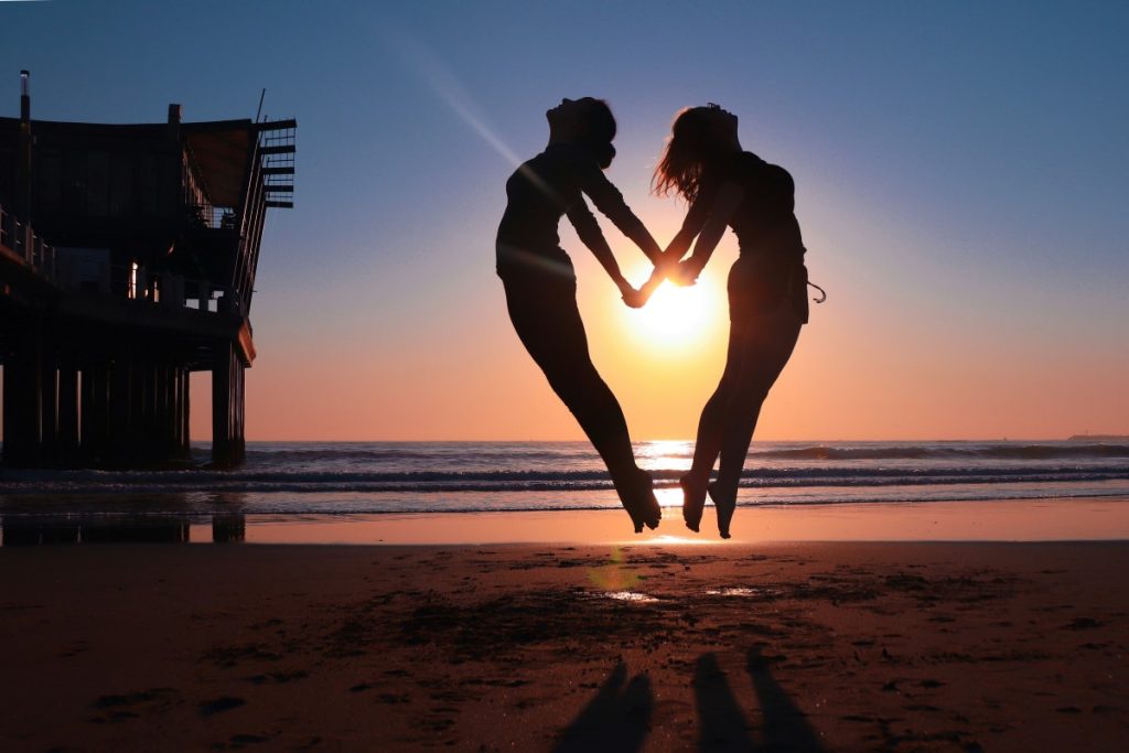 Two women jump back to back on a beach.