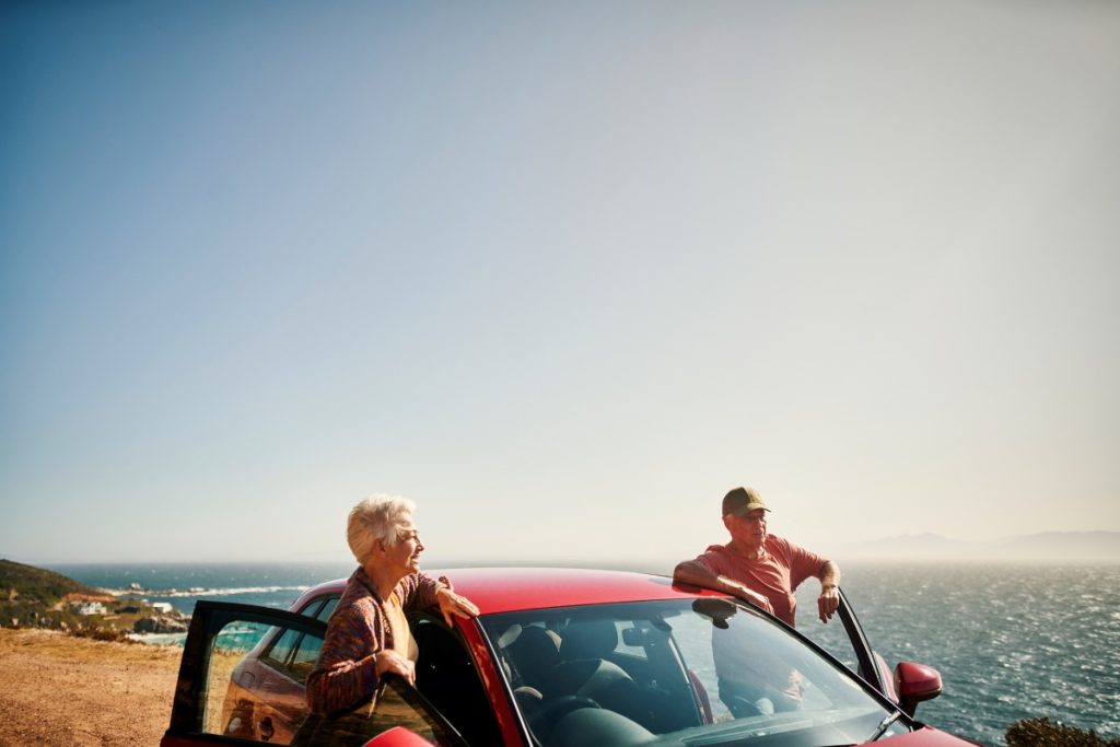 An elderly couple stands alongside their red car.