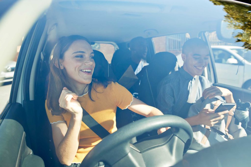 Happy young woman dancing with friends seen through car windshield on sunny day.