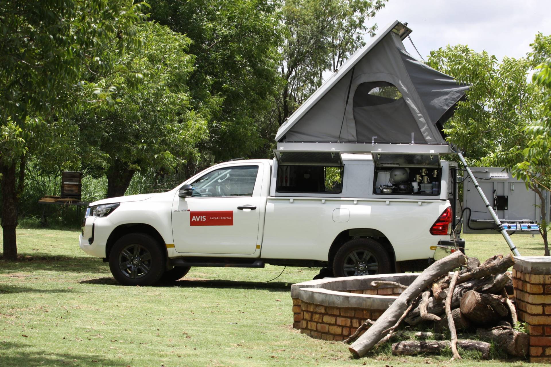 An Avis 4x4 Toyota Hilux with a rooftop tent and camping gear at a campground surrounded by trees and an outdoor firepit. 