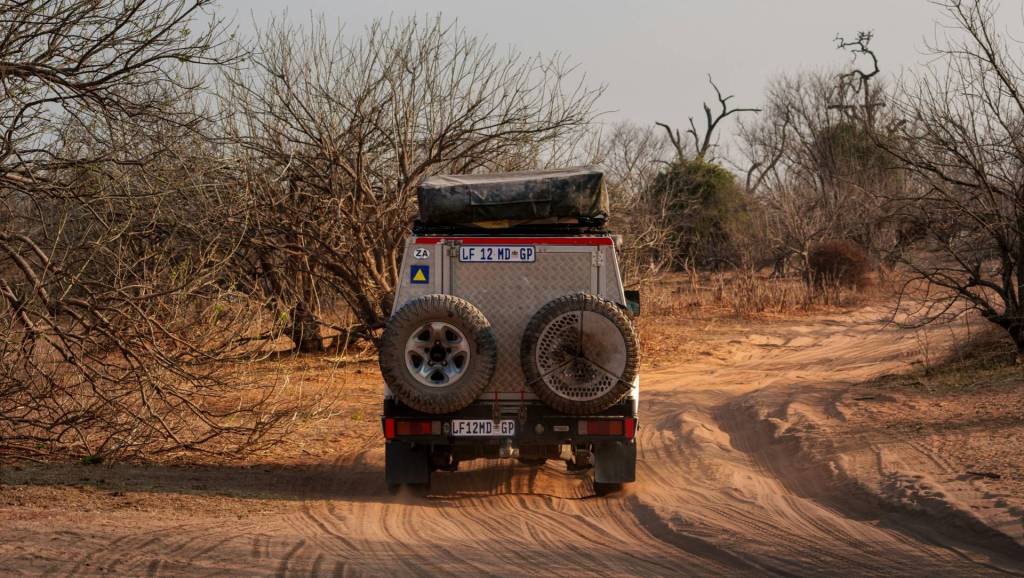A 4x4 safari vehicle equipped with gear driving along a sandy dirt road through dry bushland