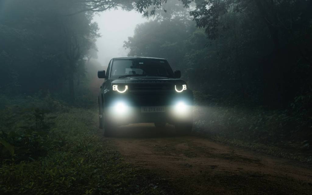 Land Rover Defender driving on an off-road track through a foggy bush area.